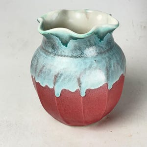 May include: A handcrafted ceramic vase with a unique design. The vase features a red base, transitioning to a blue-green middle section with a dripped glaze effect, and a ruffled, off-white rim. The vase has a textured, ribbed exterior.