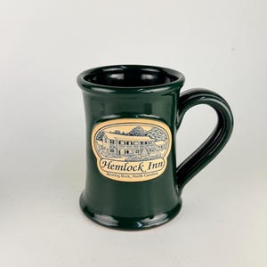 May include: A dark green ceramic mug with a glossy finish and a curved handle. An oval cream-colored decal on the front features a black line drawing of a building with the text Hemlock Inn and Blowing Rock, North Carolina printed below it.