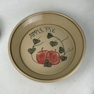 May include: A round, beige ceramic pie dish with the words "APPLE PIE" in black script. The center features a red apple design with green leaves and vines. The dish has a slightly raised edge.