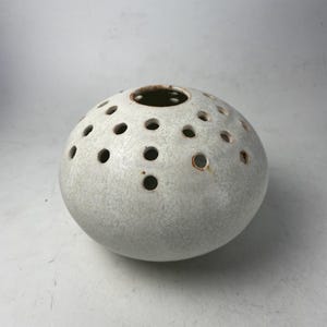 May include: A round, light gray ceramic vase with numerous small, evenly spaced holes. The vase has a textured surface and a dark brown interior visible at the top opening. The overall shape is spherical, with a slightly flattened base.