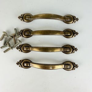 May include: Four antique brass-toned metal drawer pulls with a curved handle design and decorative ornate ends. Each pull features a smooth central bar and detailed filigree-style mounting plates. Eight silver-colored metal mounting screws are included.