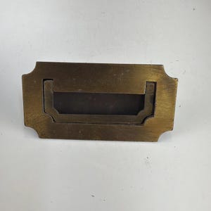 May include: A rectangular brass flush mount drawer pull with a recessed handle. The metal hardware features a weathered, aged finish with a dark patina and a notched, decorative edge design. This functional piece of furniture hardware is solid.