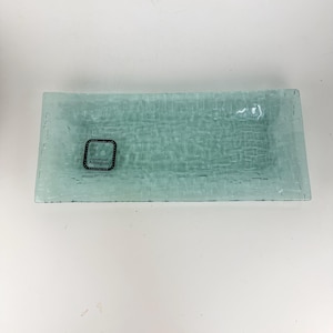 May include: A rectangular, light aqua-colored glass tray with a textured, woven-like pattern throughout the surface. A small, dark square sticker is attached to the top left corner, featuring the text "handmade Anneglass California".