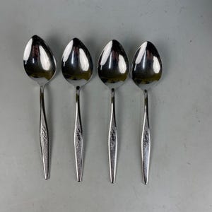 May include: Four stainless steel spoons arranged in a row. Each spoon has a teardrop-shaped bowl and a slender handle with a decorative floral pattern. The spoons are reflective, with a polished finish.