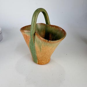 May include: A ceramic basket with a flared, asymmetrical rim and a single arched handle. The exterior features a textured finish with a blend of earthy orange and muted green glaze, showing a drip-like pattern that extends from the handle.