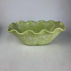 May include: A light green ceramic bowl with a ruffled edge and a white splatter pattern. The bowl has a wide, oval shape and is likely used for serving or display. The bowl is made of ceramic.