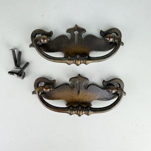 May include: Two matching antique-style brass drawer pulls with a dark, weathered patina and a central keyhole cutout. Each pull features a curved, swinging handle attached to a decorative backplate. Four small metal mounting screws are included.