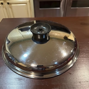 May include: A stainless steel pot lid with a black handle. The lid is round and reflective, showing the surrounding kitchen. The handle is a small, cylindrical shape. The lid is sitting on a dark brown surface.