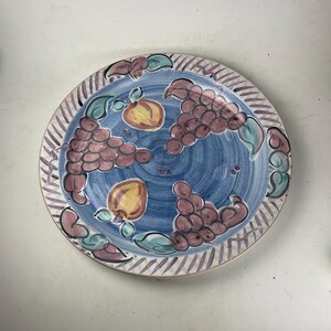 May include: A round, hand-painted ceramic plate with a blue center and a striped border. The plate features illustrations of purple grapes, yellow peaches, and green leaves. The rim has a pattern of alternating white and purple stripes.