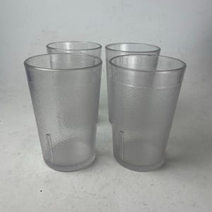 May include: Four clear, textured plastic drinking glasses. The glasses are cylindrical with a slightly flared top and a textured surface. Each glass has a vertical indentation near the base. The glasses are empty and arranged in a group.