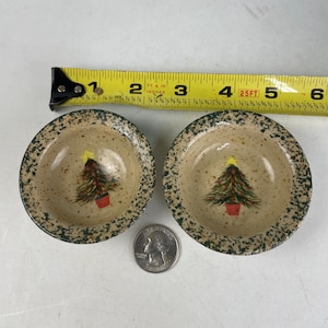 May include: Two small, round, ceramic bowls with a Christmas tree design. The bowls have a speckled tan interior and a green speckled rim. Each bowl features a hand-painted Christmas tree with a yellow star and a red pot. A tape measure shows the bowls are approximately 4 inches in diameter.