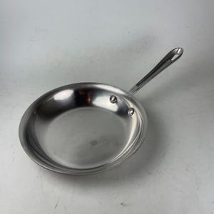 May include: A stainless steel frying pan with a long handle. The pan is round with a slightly curved edge and two rivets attaching the handle. The handle has a hole at the end for hanging. The pan is reflective.