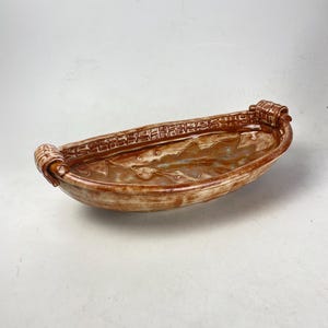 May include: A handmade ceramic serving dish with a boat-like shape. The dish is primarily brown with a textured rim and decorative handles. The interior has a glazed finish with hints of blue and white.