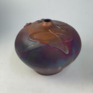 May include: A round, ceramic vase with a dark purple and bronze metallic finish. The vase features raised, stylized leaf designs and a small opening at the top. The color transitions from purple to bronze, creating a unique decorative piece.