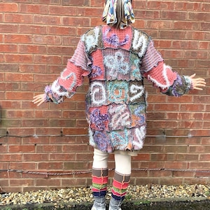 May include: A patchwork knit coat in shades of pink, purple, blue, and white. The coat features a variety of textures and patterns, with a longline silhouette. The person is wearing white leggings, white shoes, and colorful leg warmers.