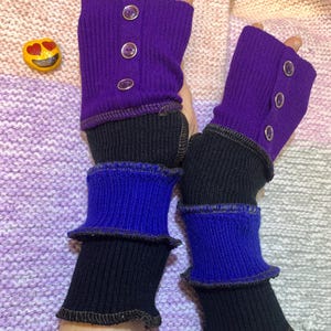 Puede incluir: Guantes sin dedos en morado, negro y azul. La parte superior es de punto morado acanalado con tres botones transparentes. La parte inferior es negra, pasando a azul con un borde con volantes. Un accesorio divertido y colorido.