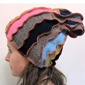 Pode incluir: Um gorro de malha patchwork colorido com bordas onduladas. O gorro apresenta seções rosa, cinza, preto e azul, costuradas com fio marrom. A borda é adornada com contas douradas, criando um design único e artístico.
