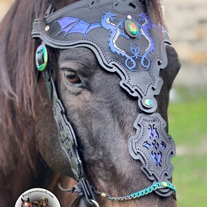 May include: A black leather horse bridle with a dragon design and iridescent blue accents. The bridle features a noseband with a decorative cutout and a browband with two dragons facing each other. The browband also has a large, iridescent green gemstone in the center.