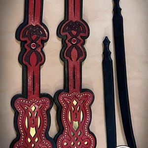 Puede incluir: Dos correas de cuero rojo con intrincados diseños de nudos celtas y detalles dorados. Las correas tienen un respaldo negro y están cosidas con hilo blanco. También se muestran dos correas de cuero negro con bordes decorativos.