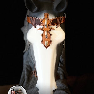 Pode incluir: Um frontal de cabeça de cavalo de couro castanho com uma cruz metálica decorativa e um design de asas de morcego. O frontal é adornado com tachas prateadas e é mostrado em uma cabeça de cavalo branca.
