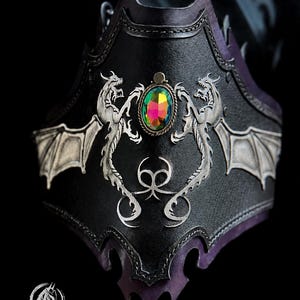 May include: A black leather bracer featuring two silver-toned dragon emblems with spread wings flanking a central oval iridescent gemstone. The piece includes decorative stitching along the edges and a logo reading FANGORN RANCH at the bottom.