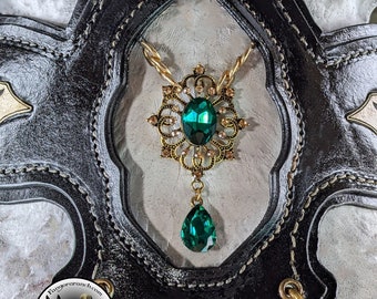 Könnte beinhalten: Eine goldene Halskette mit einem grünen Edelstein-Anhänger hängt an einem schwarzen Ledergeschirr. Der Anhänger hat ein detailliertes Filigran-Design mit kleineren klaren Edelsteinen.