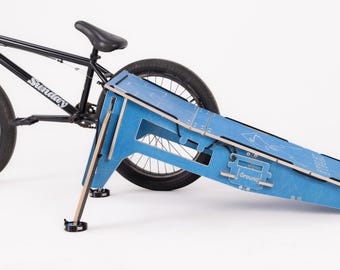 Può includere: Una bici BMX nera accanto a una rampa di legno blu. La rampa è dipinta di blu con bordi in legno naturale e la parola "Ground" è visibile. La bici ha la scritta "Sunday" sul telaio.