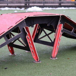 May include: A red and black wooden ramp with a flat top and angled sides. The ramp is made of plywood and has a black frame. The ramp is sitting on a green artificial grass surface.