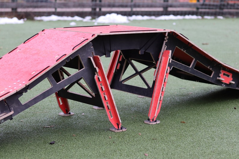 May include: A red and black wooden ramp with a flat top and angled sides. The ramp is made of plywood and has a black frame. The ramp is sitting on a green artificial grass surface.