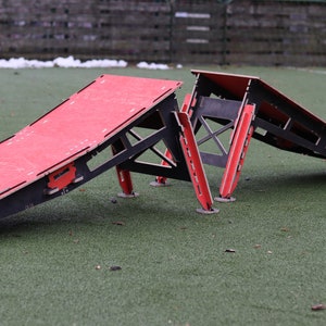 May include: Two red and black wooden ramps for skateboarding or other action sports. The ramps are set up on a green artificial turf surface.