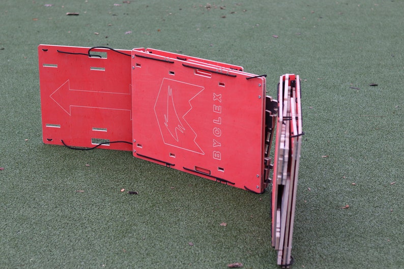 May include: A red, foldable, portable agility hurdle with the word "BYCLEX" printed on it. The hurdle is made of wood and has a black strap.