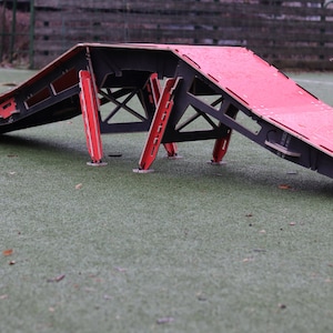 May include: A red and black wooden ramp with a triangular shape and four legs. The ramp is made of plywood and has a textured surface.