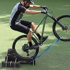 Peut inclure: Une personne fait du vélo sur un support d'entraînement. Le vélo est vert, la roue avant est levée et une sangle bleue relie la roue au support. Le support porte le texte "RYCLEX".