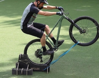 May include: A person riding a green mountain bike on a training stand. The bike's front wheel is raised, and a blue strap connects the wheel to the stand. The stand has the text "RYCLEX" on it.