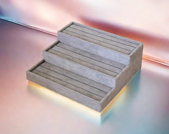 May include: A gray tiered ring holder with three levels. Each level has a soft, gray felt lining to protect rings.