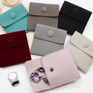 May include: A selection of small, colourful velvet pouches in teal, grey, black, burgundy, light grey, and cream. One pink pouch is open, displaying a bracelet. A silver ring with a dark blue stone is also visible.