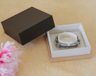 May include: A silver bracelet with a detailed design and clear stones is displayed in a white velvet lined wooden box with a black lid.