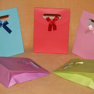 May include: Five gift bags in different colors, each with a bow. The bags are light blue, red, pink, purple, and green. The bags have a cut-out handle and are made of paper.