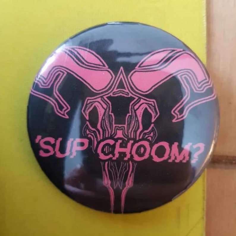 May include: A black button with a pink graphic of a stylized creature with the text 'SUP CHOOM?'