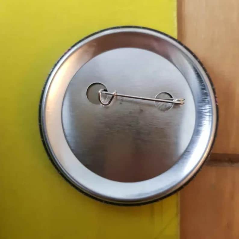 May include: The back of a round metal button with a silver finish. The button has a pin back with a safety clasp.