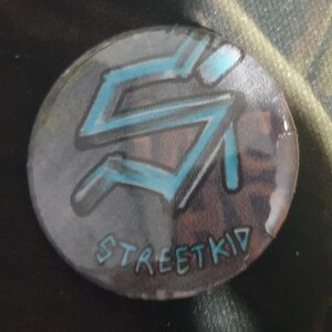 May include: A round, blue and white button with the word "STREETKID" and a stylized letter "S" design.