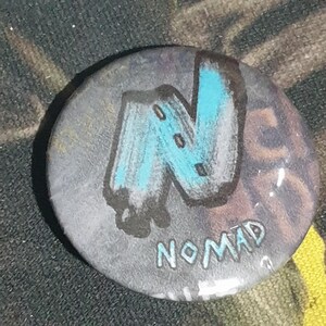 May include: A round, gray button with a blue and black stylized letter "N" and the word "NOMAD" written below it.
