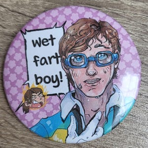 May include: A circular badge featuring a cartoon illustration of a person wearing blue glasses and a blue and white shirt. The badge has the text "wet fart boy!" in a speech bubble. The background is light purple with a pink floral pattern.