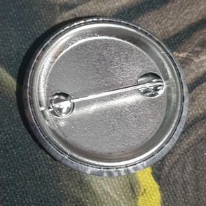May include: The back of a silver button with a pin and a safety clasp. The button is round and has a smooth surface.