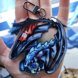 May include: A clear acrylic keychain featuring a stylized dragon design. The dragon is primarily blue and black with red accents on the wings. The keychain has a silver-colored clasp for attachment.