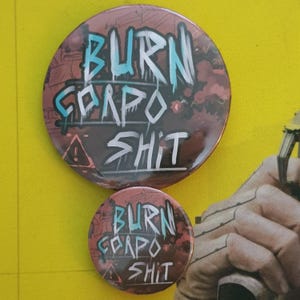 Puede incluir: Dos botones redondos con un diseño gráfico de estilo grunge. Los botones son rojos, negros y blancos con el texto "BURN CORPO SHIT" en letras azules y blancas.