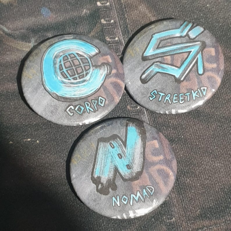 May include: Three round, gray, pinback buttons with blue and white painted designs. The first button says "CORPO" and has a blue circle with a white grid inside. The second button says "STREETKID" and has a blue, stylized letter "S". The third button says "NOMAD" and has a blue, stylized letter "N".