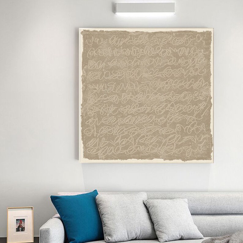 Puede incluir: Un arte de pared de lienzo beige con un dise&ntilde;o abstracto blanco. El arte est&aacute; colgado en una pared blanca sobre un sof&aacute; gris claro con almohadas azules y beige.