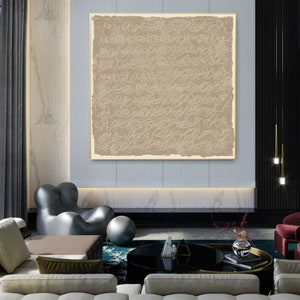 Puede incluir: Un arte de pared de lienzo beige con un dise&ntilde;o abstracto blanco. El arte est&aacute; colgado en una sala de estar moderna con un sof&aacute; gris, una mesa de caf&eacute; negra y una alfombra gris y blanca.