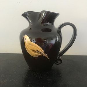 Puede incluir: Una jarra de cerámica negra con un pájaro amarillo pintado en el lateral. La jarra tiene una boca ancha y un asa.